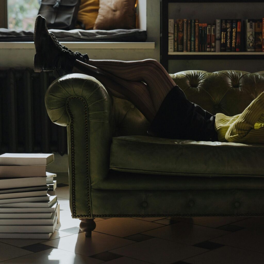 Mujer acostada en el sofá con leyendo un libro,conlospies sobre el apoyabrazos, una biblioteca de fondo y más libros al pie del sofá. Seguro está leyendo como escritora.
